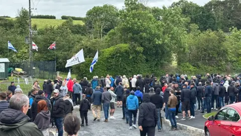 PA A large number of people are standing on a road. They are facing a wooded area. Five flags can be seen flying above them. Two blue and white flags, one red and white flag, one red white and blue flag, and a white flag.