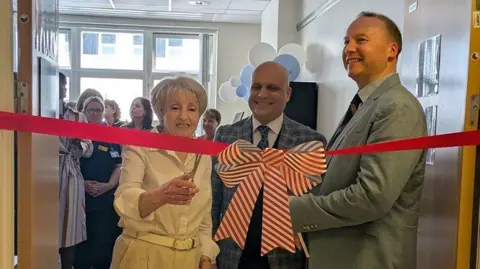 Sylvia Wyer is wearing a white blouse and beige trousers, and she has short white hair. She is holding a pair of scissors and is about to cut a large red ribbon stretched across a doorway, with a white and red striped bow. Next to her is a man in a striped suit jacket. There is another man next to him in a grey suit