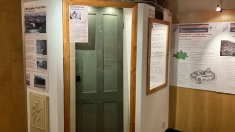 A green painted wood door with glass panels inset stands behind a transparent plastic case. Either side of it are white painted walls with exhibition information pinned to them.