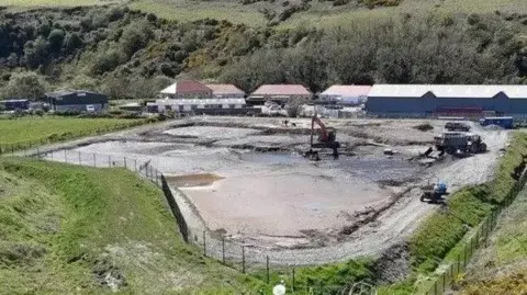 IOM GOVERNMENT Picture of the silt lagoon. An elevated square area of field can be seen with work sheds behind and treatment facility works next to it.