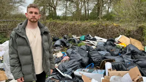 A man wearing a green puffer jacket. He has ginger hair and is standing in front of a pile of rubbish