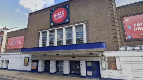 Google A brick building with some boarded up windows and some white painted walls and pillars. There are several red signs, including one which say" Buzz Bingo".