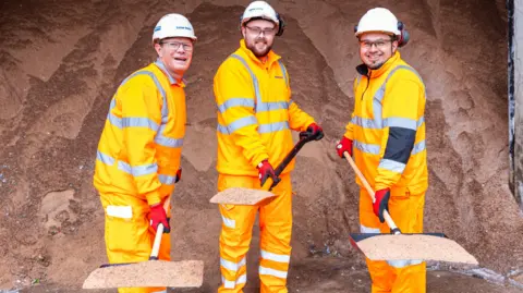 Three men in orange hi-vis protective clothing, white hard harts and protective goggles, who are holding shovels full of grit, and standing in front of a bigger pile of grit.