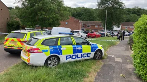 Two police cars block the road. One is positioned on the grass bank while another is parked in the road. Police tape marks the start of the cordon and three men are seen in a group further down the path in the cordon. 