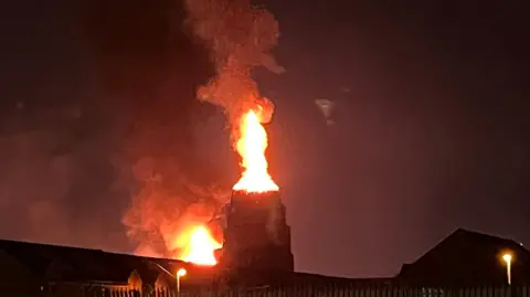 Flames are coming out of a tower, silhouetted against a dark sky. You can see some rooftops and street lights.