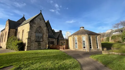 A path leading to the oval-shaped cafe, which is adjacent to the church. Both building are made of beige stone. 