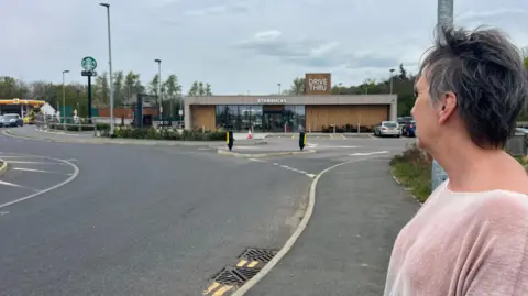 BBC/Owen Sennitt A woman wearing a pink top looks towards a roadside services on a grey spring day