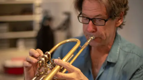 John Wallace playing a trumpet. He has the instrument to his mouth and is blowing on the mouthpiece. He is wearing black-rimmed glasses and has floppy hair. He is wearing a denim shirt. 