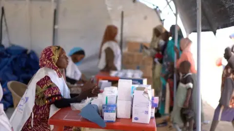 Reuters A medic waits in a makeshift clinic as displaced Sudanese gather. There is a large pile of white boxes in front of her.