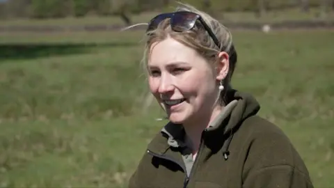 Cara Hewitt, a ranger at Bradgate Park. She has blonde hair, is smiling and wearing a green fleece jacket. She has sunglasses on her head. In the background is a green field.