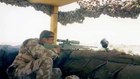 Ross family Michael Ross wearing Army fatigues looking through the sight of a rife in a dugout made of sand bags. 