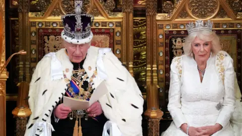 The King and Queen are sitting in large gold chairs. They are both wearing crowns. King Charles is addressing MPs and is reading a speech from a book in his hands. 