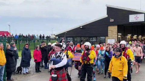 Swimmers in various outfits march out of the lifeboat station behind a bagpiper in traditional Scottish dress