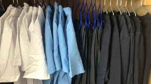 Getty Images White and light blue polo shirts are hung up on hangers on a rail to the left. On the right are trousers and black jumpers hung up on the same rail.