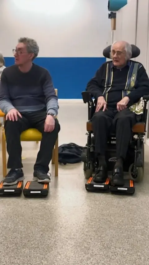 Two elderly men sat in chairs with pedal devices at feet
