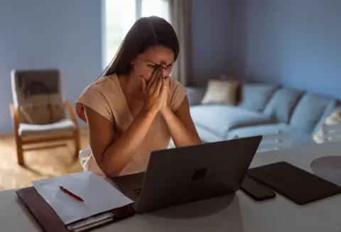 A woman sits at a desk looking at her laptop screen with her head in her hands