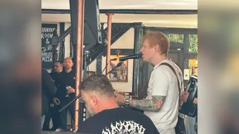 Richard Knights/BBC Ed Sheeran in a pub, playing his guitar and singing. Other musicians members can be seen. people mill about holding drinks.