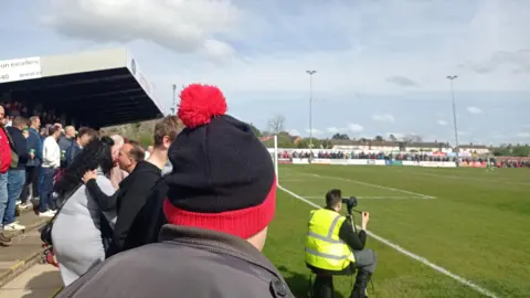 Nireas Habidis The back of the head of a man who is wearing a black and red bobble hat. He is standing on the terraces among hundreds of other people during a match at Kettering Town Football Club.
