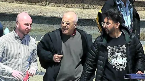 Reece McLaren, Terence O’Hagan and Shahiukh Syed Gilani are standing together in front of a building on a sunny day. McLaren has a bald head and is wearing a striped shirt. O'Hagan is also bald and is wearing a grey sweatshirt and black coat. Gilani has dark wavy hair and is wearing a black sweatshirt that has a tiger face traced in white under a black puffer jacket, and is holding a pizza box. 