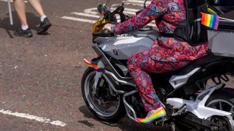 PA Media A Pride parade participant rides a motorcycle decorated with small rainbow flags while wearing a brightly patterned paisley suit and rainbow-coloured high-heeled shoes.