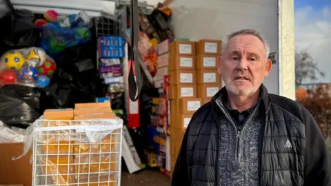 LDRS David Taylor with short grey hair and beard wearing a grey zip up jumper and black jacket. He stands in front of a white van where he has put his stock from his toy stall.
