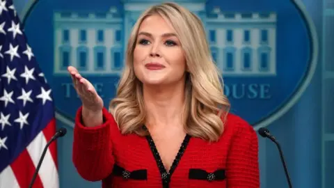 Karoline Leavitt is behind a podium in the white house briefing room holding her right hand palm facing up.