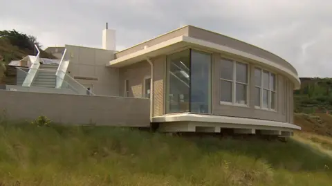 A wide shot of the empty former restaurant building. It is built on the seafront. Grassland surrounds the property which has large windows.