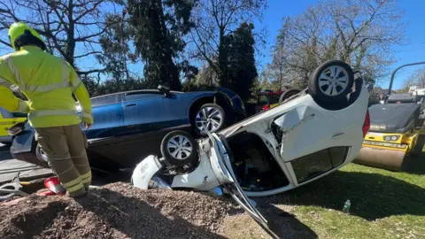 Essex Fire and Rescue Service The scene of a car crash. A blue Volkswagen is resting on the underside of a white Hyundai, which has crashed on its roof and is wedged into the ground. Trees are in the background, while a firefighter is standing on the left.