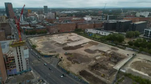 EPA An aerial photograph taken with a drone shows construction on a large, brownfield site in Manchester. Tower blocks can be seen in the distance, smaller office blocks are nearby and there is a multi-lane road running along the left hand side. A red crane stands on the left hand side of the picture.