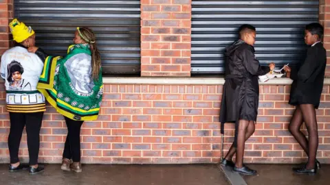 Getty Images Two schoolgirls have a snack as two women wearing African National Congress (ANC) regalia pause at the Orlando stadium in Soweto, Johannesburg, on April 11, 2018.