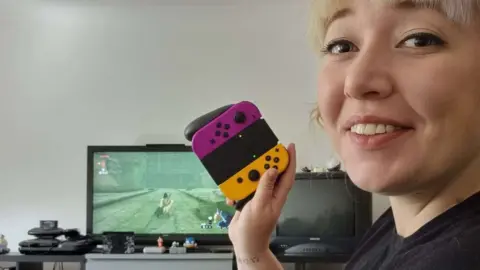 Sky Sky is at the right-hand side of the frame, smiling. She's holding up a Nintendo Switch controller, and in the background a TV, surrounded by different games consoles, is playing a Zelda game