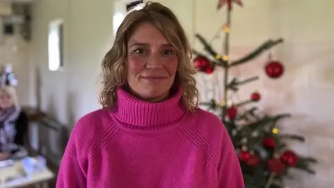 Jasmine Watson. She is stood in front of a Christmas tree. She has blonde hair and is wearing a bright pink jumper. She is smiling at the camera.