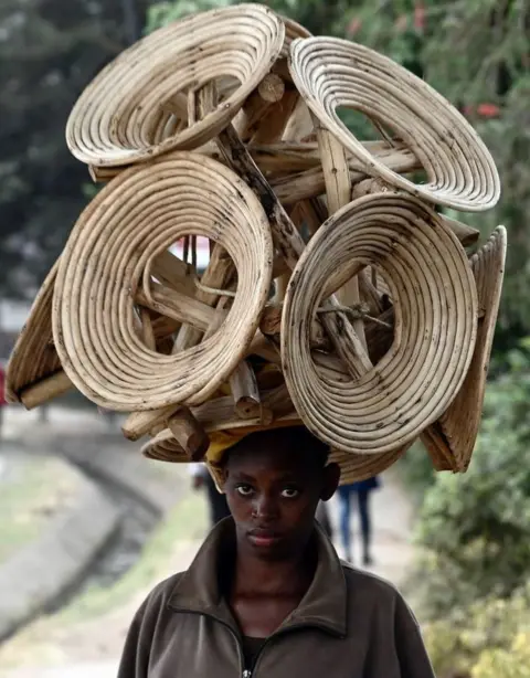 AFP A woman carries several wooden chairs on her head on a street. She is looking directly at the camera.