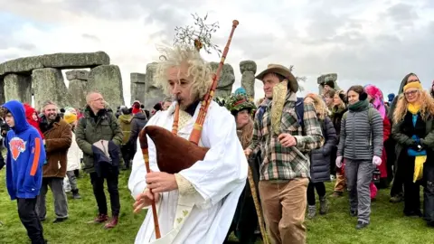 Ben Birchall/PA Crowds at Stonehenge including a man wearing a white robe while playing the bag pipes