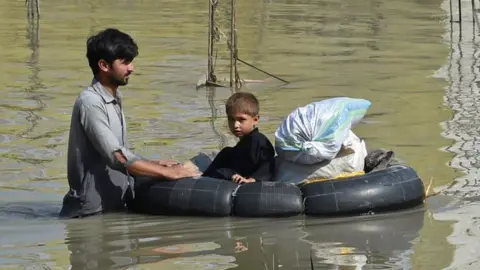 Getty Images A man with child wades through a flood hit area following heavy monsoon rains in Charsadda district of Khyber Pakhtunkhwa.