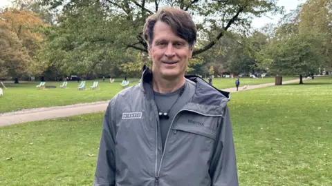 A middle-aged man with brown hair, parted at the side, stands in a park and smiles. He's wearing a grey t-shit underneath a thin, grey waterproof jacket which has a "Niantic" logo on the right breast and a "Marmot" logo on the left. He's got a pair of earbuds joined by a wire hanging from his neck. Behind him, we can see deck chairs, trees with green leaves just starting to show the first signs of autumn, and a woman walking her dog on a path that cuts through the park's grassed area.