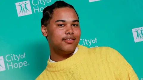 Getty Images Singer songwriter Sekou Sylla pictured at an event in 2022. Sekou is a 19-year-old man with short black hair which he has braided. He has a small moustache and brown eyes and smiles slightly at the camera. He wears a yellow knitted jumper over a white shirt.