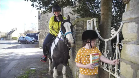 PA Media Rescue pony Micky helps owner Abi Eliot-Williams and her daughter Polly, deliver library books to members of the Hullavington Book Group in the village of Hullavington near Malmesbury, Wiltshire.