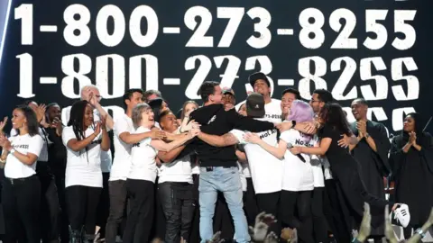 Getty Images Logic hugs survivors of suicide on stage, with the helpline number clearly visible on shirts and screen behind
