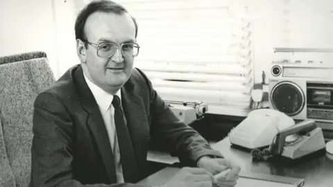 Paul Deal Black and white photo of Paul Deal in 1984, wearing glasses and sat at a desk beside two phones and a radio