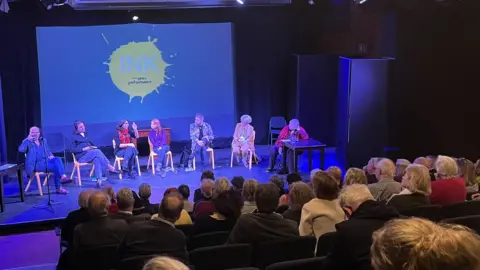 Jon Wright/BBC Seven people on chairs on a stage in front of an audience in a theatre