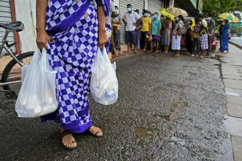 Getty Images Woman carrying food bags in Colombo while people wait in queue outside a state-run supermarket