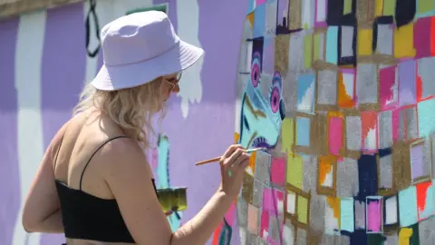 Georgie Webster Georgie Webster wearing a lilac hat painting a wall in colourful squares