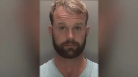 Man with brown hair and beard wearing light-coloured shirt in police mugshot