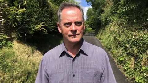 BBC Mike Phillips looking at camera while standing on road with hedgerows either side