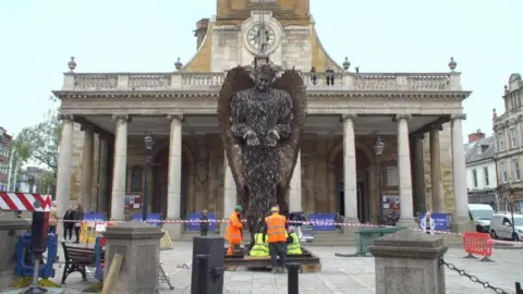 Stuart Ratcliffe/BBC Human figure made up of knives in front of stone-pillared All Saints Church piazza