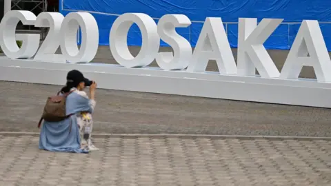 Getty Images A woman takes a picture of a G20 Osaka design set up outside the venue for the G20 Osaka Summit in Osaka on June 26, 2019, ahead of the start of the summit later this week.