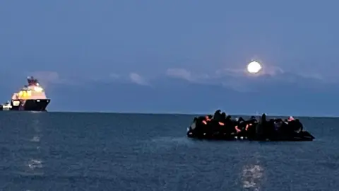 BBC News An overcrowded inflatable boat heads out to sea in the English Channel under moonlight. A French coastguard ship is on the horizon.