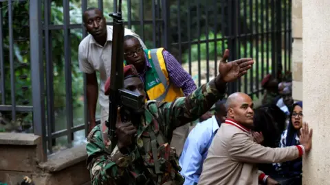 Ronald Grant A member of the security forces gestures as people take cover at the scene where explosions and gunshots were heard at the Dusit hotel compound, in Nairobi, Kenya