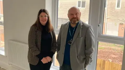 LDRS a woman and a man standing in front of a window in an empty room in a newly-built home with blank walls and uncovered floors